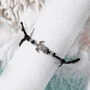 Detail of silver turtle charm on black braided cord anklet with beach shells backdrop