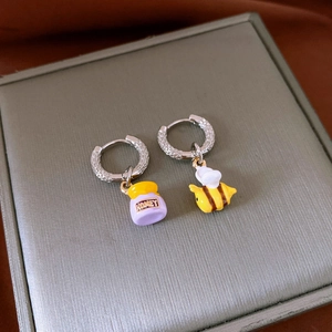 Detail of bee and purple honey jar charms on silver hoop earrings over a grey gift box
