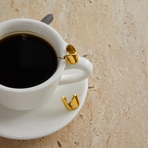 Polished gold organic C-shape hoop earrings in stainless steel on white coffee cup