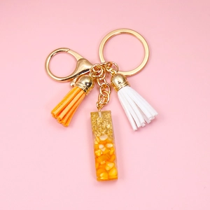 Detail of gold keychain with orange tassel and corn kernel resin charm on pink background