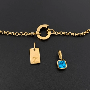 Detalle de un collar de acero de titanio dorado con placa de la letra Z y dije de circón azul