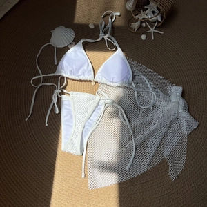 Flat lay of white shell bikini 3-piece set with mesh cover-up on a textured mat