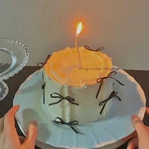 Close-up of white cream cake with brown bows and a burning candle held by hands