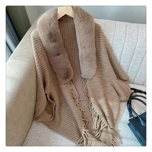Detail of beige knit poncho with faux fur collar and fringe trim on a textured fabric