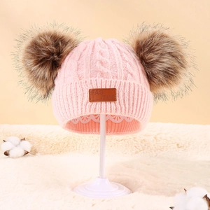 Close-up of pink cable knit baby hat with dual brown faux fur pom poms and leather patch
