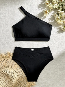 Flat lay of black ribbed one-shoulder bikini set with high-waisted bottoms on woven mat