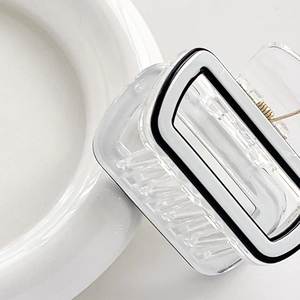 Pince à cheveux carrée transparente avec contours noirs et ressort métallique sur plat blanc