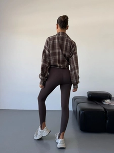 Brown plaid long sleeve shirt with dark leggings and white sneakers in a minimalist room