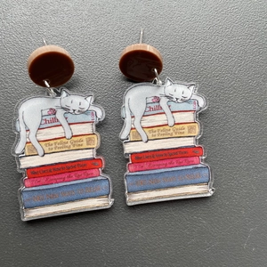 Detail of acrylic earring showing a white cat sleeping on books with Chill text titles