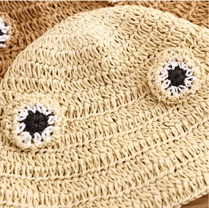 Close-up of beige woven straw hat featuring cartoon frog eye decorations and texture