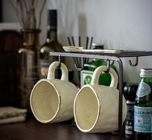 Brown metal coffee mug rack with hooks holding beige handmade ceramic mugs on top shelf