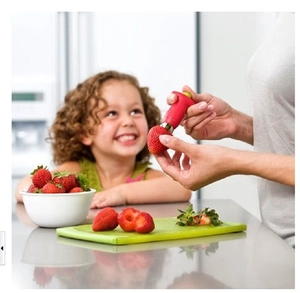 A person using the red strawberry huller tool in a kitchen setting with a young girl