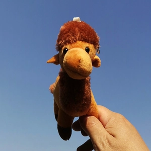 Cute brown camel plush keychain charm being held against a clear blue sky background.