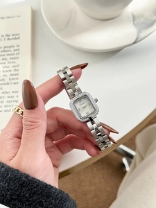 Close-up of square silver watch with crystal bezel and blue stone crown on a lifestyle backdrop