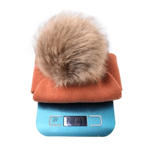 Orange knitted baby beanie with removable fur pom pom shown on a scale weighing 107g