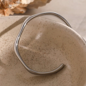 Detailed view of wave-shaped polished stainless steel open bangle on a ceramic surface