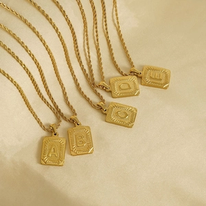 Détail de chaînes torsadées dorées 18K avec pendentifs carrés lettres A-E sur tissu beige