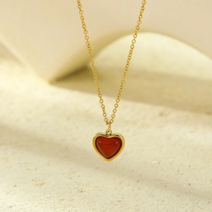Golden heart necklace with red agate stone set against a minimalist sandy background