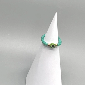 Detail of turquoise beaded stretch ring with green evil eye charm on a white stand