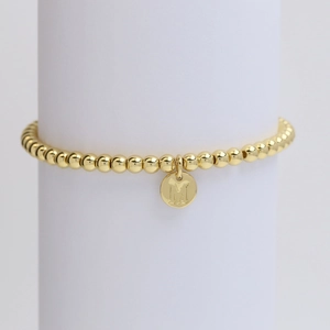 Detailed view of golden beaded bracelet with M-letter round charm on white stand