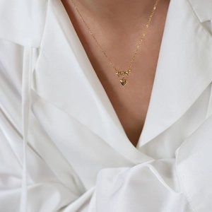 Close-up of gold bow and heart pendant necklace worn on a white silk shirt
