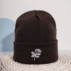 Deep brown knit beanie with white rose embroidery on a textured straw mat background