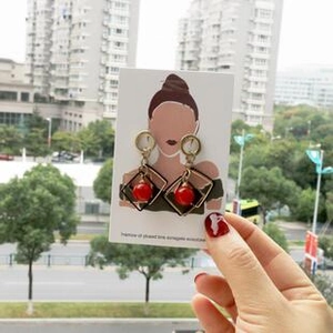 Close-up of golden geometric square earrings with red bead drops held on a display card