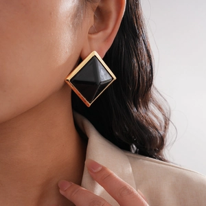 Close-up of model wearing large geometric gold-rimmed diamond-shaped earrings with black resin