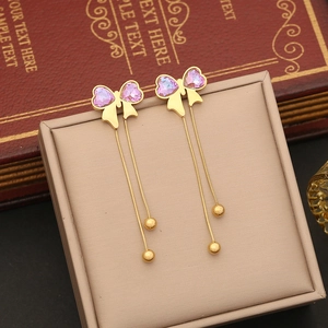 Elegant gold bow earrings with purple heart crystals and long tassel chains on a display box