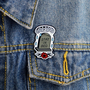 Artistic tombstone enamel pin with Stay In Bed text on a blue denim jacket