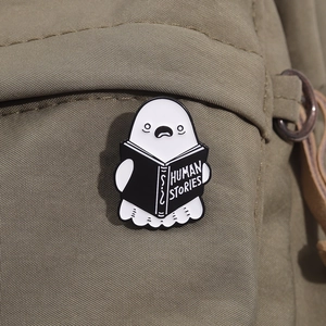 Close-up of cute ghost enamel pin reading a book on an olive green canvas backpack