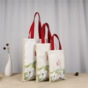 Three sizes of canvas tote bags with green landscape art and red handles on a table