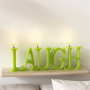 Green 'LAUGH' word candle with burning wicks on a wooden shelf for cozy home decor