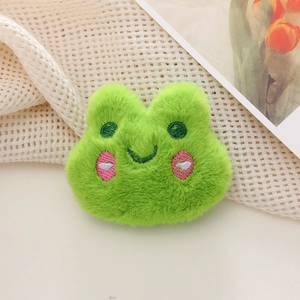 Close-up of a green plush frog brooch with embroidered eyes and pink heart cheeks