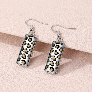 Silver-tone rectangular leopard print earrings displayed on a pink textured background
