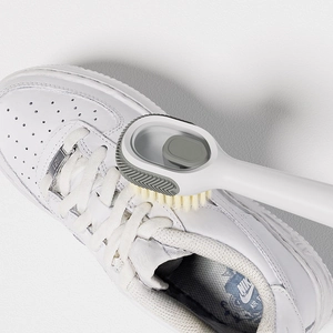 White liquid-dispensing shoe brush with soft grey bristles cleaning a leather sneaker