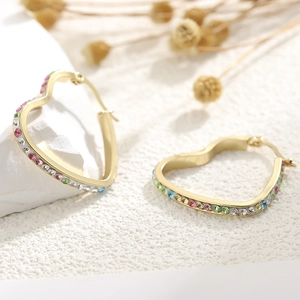 Exaggerated golden heart hoop earrings with colorful crystals on a textured white pedestal