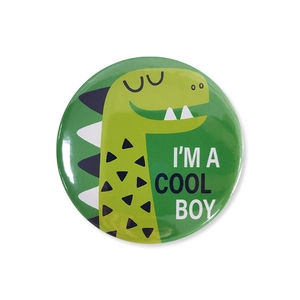 Close-up of round tinplate badge featuring a green cartoon dinosaur and I'M A COOL BOY text