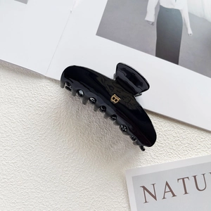 Detail shot of a glossy black hair claw clip featuring a metallic gold letter B logo