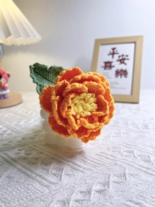 Handmade crochet peony flower with orange petals and green leaves on a white stand