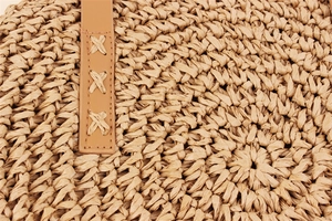 Detailed close-up of woven paper rope texture and brown leather stitching on a round beach bag