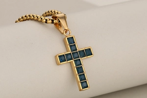 Close-up of golden cross pendant with blue square gemstone on a beaded chain necklace