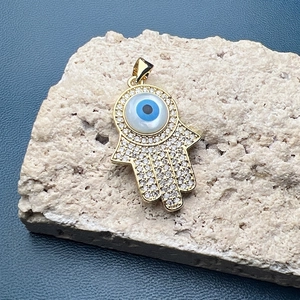 Detailed view of a gold Hamsa hand pendant with pavé zircons and a blue evil eye bead