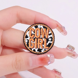 Cow Girl enamel brooch with orange lettering and cow print, held in hand display
