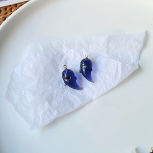 Close-up of a pair of translucent dark blue ghost charms with golden metal loops on white paper
