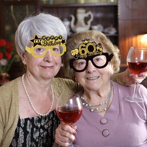 Two happy senior women wearing black and gold 60th birthday party paper glasses with glitter