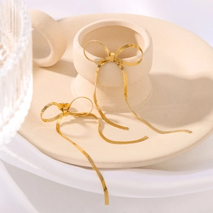 Golden stainless steel bow tassel earrings with snake chain detail on ceramic stand