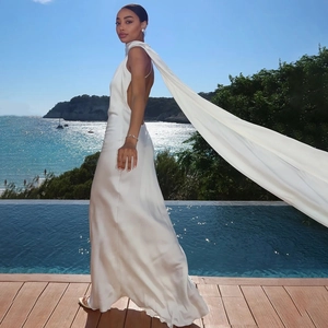 Elegant white backless maxi dress with long flowing cape detail against an ocean backdrop