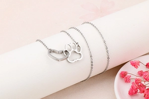 Close-up of a silver heart and cat paw pendant on a delicate chain over pink background