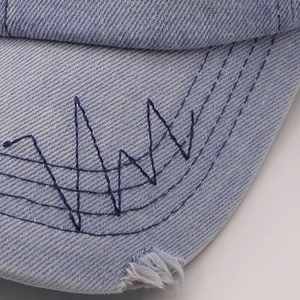 Detail of light blue washed denim baseball cap with navy embroidery and distressed brim